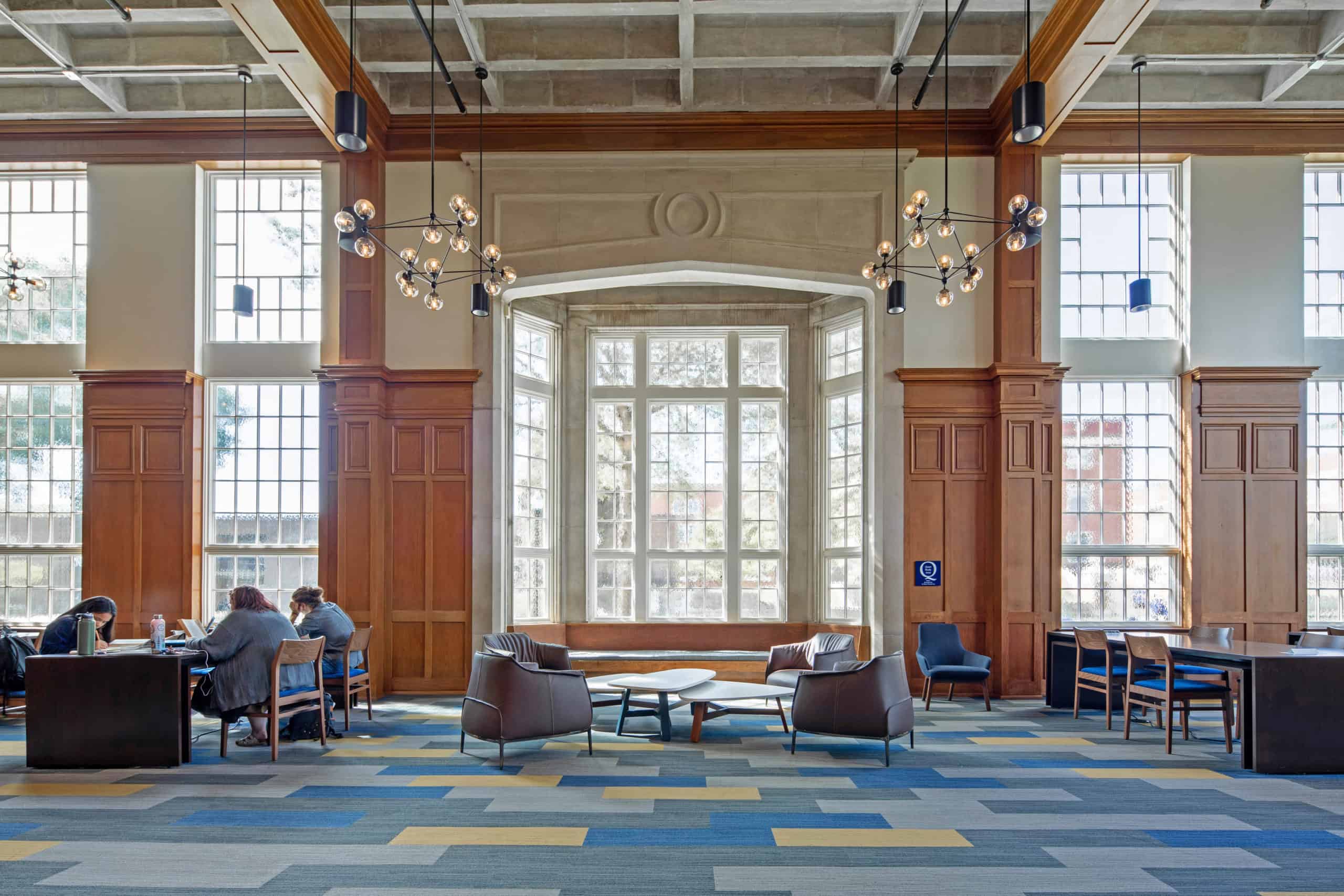 A large room with traditional, carved wood wall paneling, concrete casted walls, modern furniture and light fixtures, stained glass, and students working. This photo is to demonstrate the preservation and modernization that HK Architects implemented in the Guerry Center at the University of Tennessee at Chattanooga. 